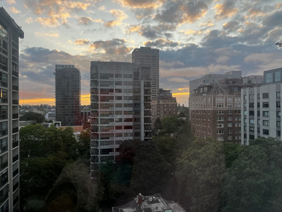 View of a city skyline at dawn, with high-rise apartment buildings in the foreground, trees below, and a partly cloudy sky glowing with orange and pink light at the horizon.